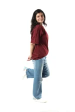 Side view of a woman in maroon t-shirt and jeans, lifting one leg playfully.